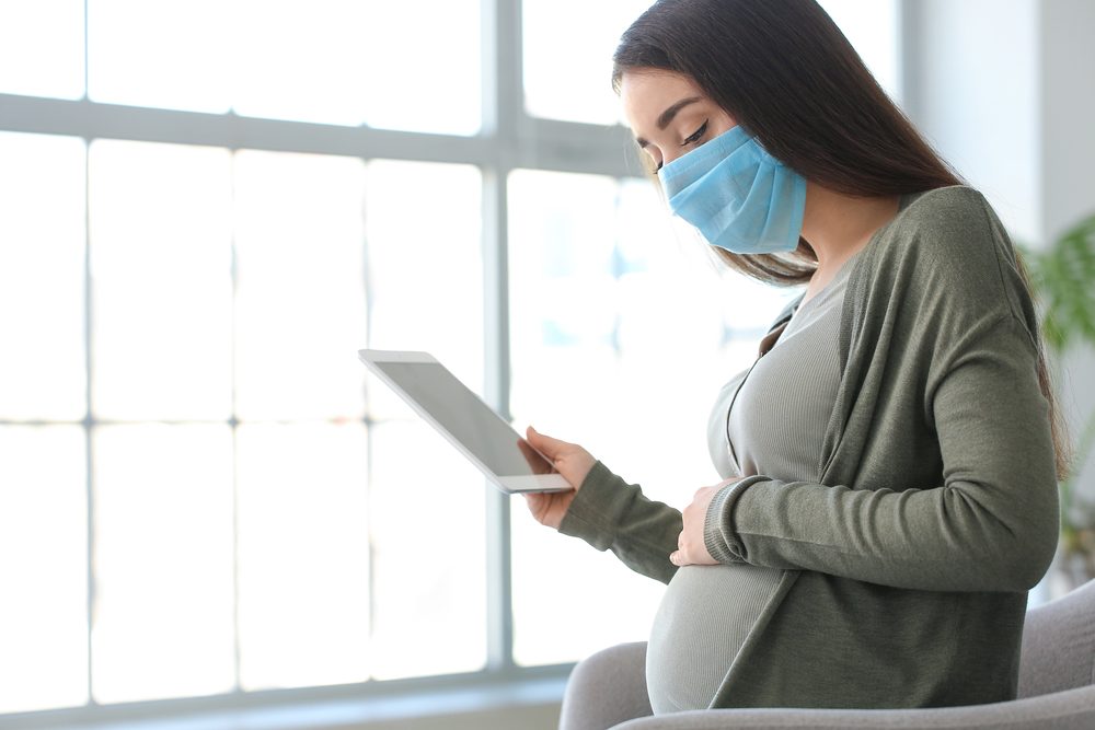 Pregnant woman with tablet computer wearing medical mask at home