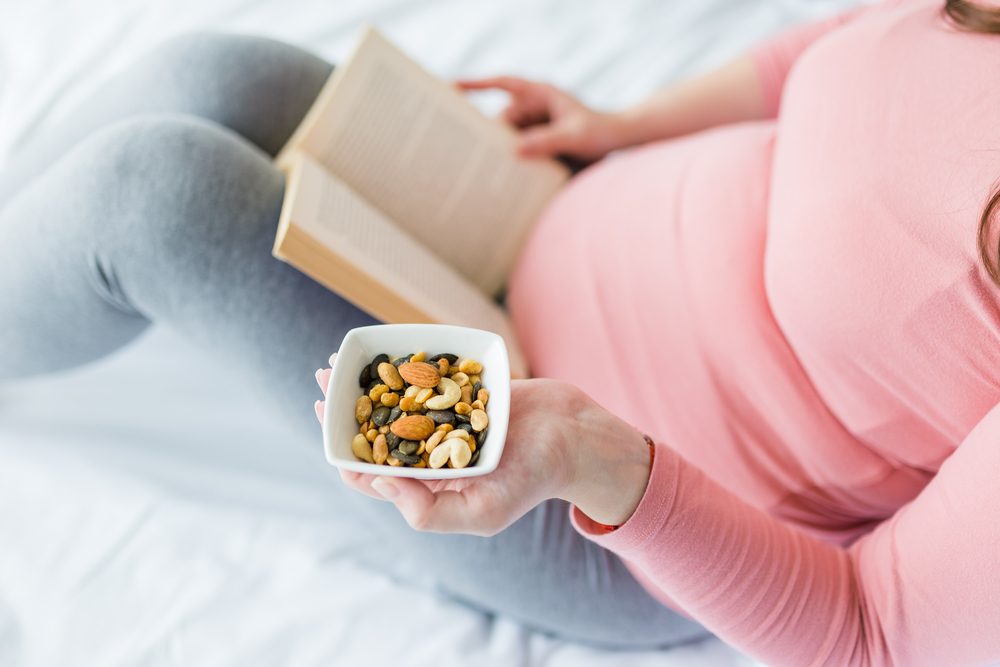 Female holding bowl with fresh nuts on bed and reading book. Pregnancy and healthy lifestyle concept.